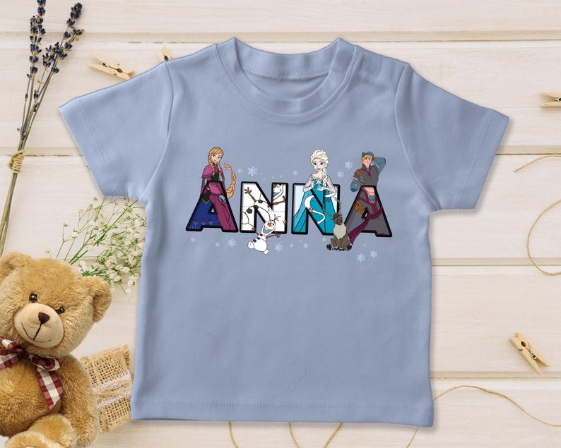 Puede incluir: Una camiseta azul claro con el nombre "ANNA" impreso en letras grandes y coloridas. Cada letra est&aacute; decorada con personajes de la pel&iacute;cula Frozen, incluyendo a Anna, Elsa, Kristoff y Olaf. La camiseta est&aacute; hecha de un material suave.