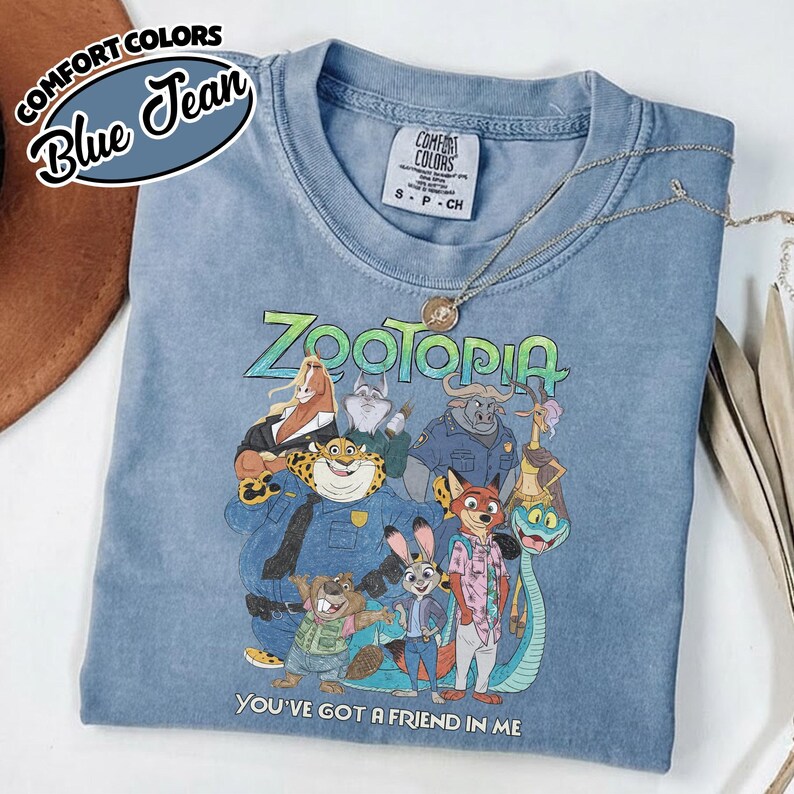 May include: Blue jean Comfort Colors t-shirt featuring a colorful graphic of characters from the movie Zootopia. The text "ZOOTOPIA" is across the top, and "YOU'VE GOT A FRIEND IN ME" is at the bottom. A silver necklace is draped across the shirt.
