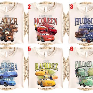 May include: Cream-coloured t-shirts featuring characters from the animated film "Cars." Each shirt displays a different character's name and image: Mater, Lightning McQueen, Doc Hudson, Sally Carrera, Cruz Ramirez, and Fillmore. The designs include illustrations of the cars.