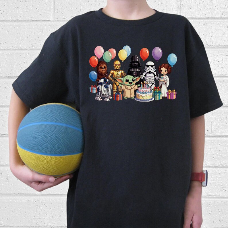 K&ouml;nnte beinhalten: Ein marineblaues T-Shirt mit einem farbenfrohen Motiv von Star Wars-Charakteren, die einen Geburtstag feiern. Das Design umfasst Yoda, Darth Vader und R2-D2 sowie Luftballons und eine Geburtstagstorte. Ein Kind h&auml;lt einen blau-gelben Basketball.