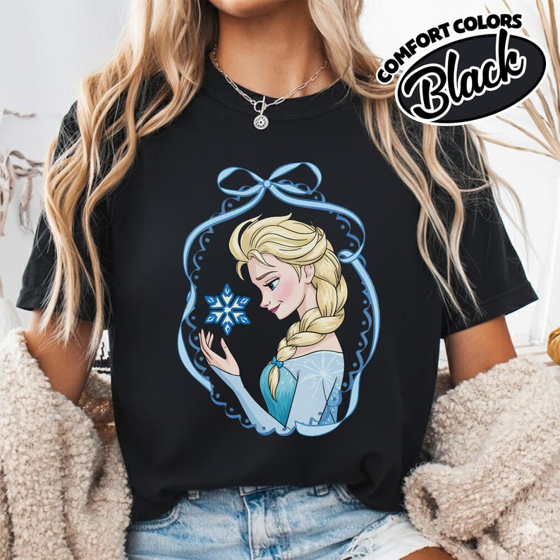 May include: Black t-shirt featuring a cartoon illustration of a blonde woman with a braid, holding a snowflake. The design is framed by a blue decorative border. The words "COMFORT COLORS" and "Black" are printed on the upper right.