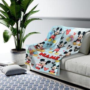 May include: A light blue fleece throw blanket featuring a repeating pattern of Mickey Mouse in various poses, colorful balloons, and the word NAME in bold red capital letters. The blanket is draped over a sofa, showcasing the cartoon character print.