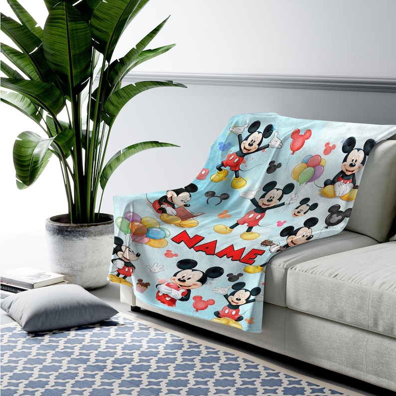 May include: A light blue fleece throw blanket featuring a repeating pattern of Mickey Mouse in various poses, colorful balloons, and the word NAME in bold red capital letters. The blanket is draped over a sofa, showcasing the cartoon character print.