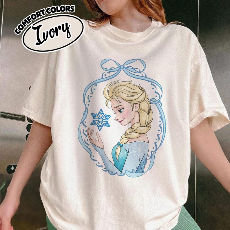 May include: Ivory Comfort Colors t-shirt featuring a cartoon illustration of a blonde woman with a blue dress and a snowflake. The illustration is framed by a light blue decorative border with a bow at the top. The words "COMFORT COLORS" and "Ivory" are printed on the top left.