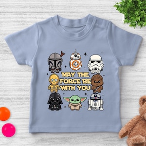 May include: Light blue t-shirt featuring cartoon illustrations of Star Wars characters, including The Mandalorian, BB-8, and Darth Vader. The phrase "MAY THE FORCE BE WITH YOU" is printed in the center of the shirt.