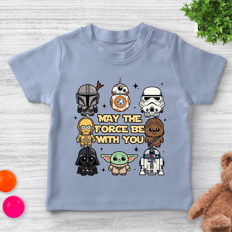 May include: Light blue t-shirt featuring cartoon illustrations of Star Wars characters, including The Mandalorian, BB-8, and Darth Vader. The phrase "MAY THE FORCE BE WITH YOU" is printed in the center of the shirt.