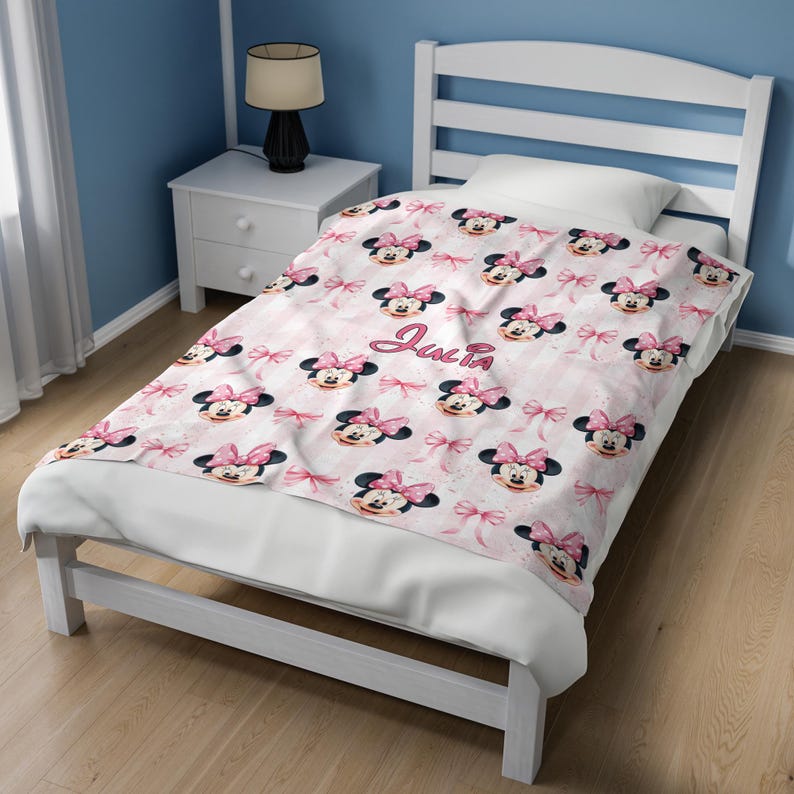 May include: A soft blanket featuring a repeating pattern of Minnie Mouse faces with pink bows and small pink bows on a light pink and white striped background. The name Julia is printed in a cursive pink script in the center of the blanket.