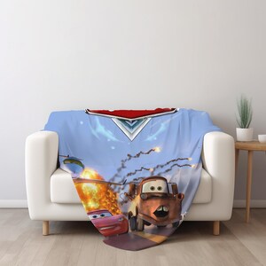 May include: A soft throw blanket featuring a vibrant graphic print of characters from the movie Cars, including a red race car and a brown tow truck, set against a light blue background with an explosion and a metallic emblem at the top.