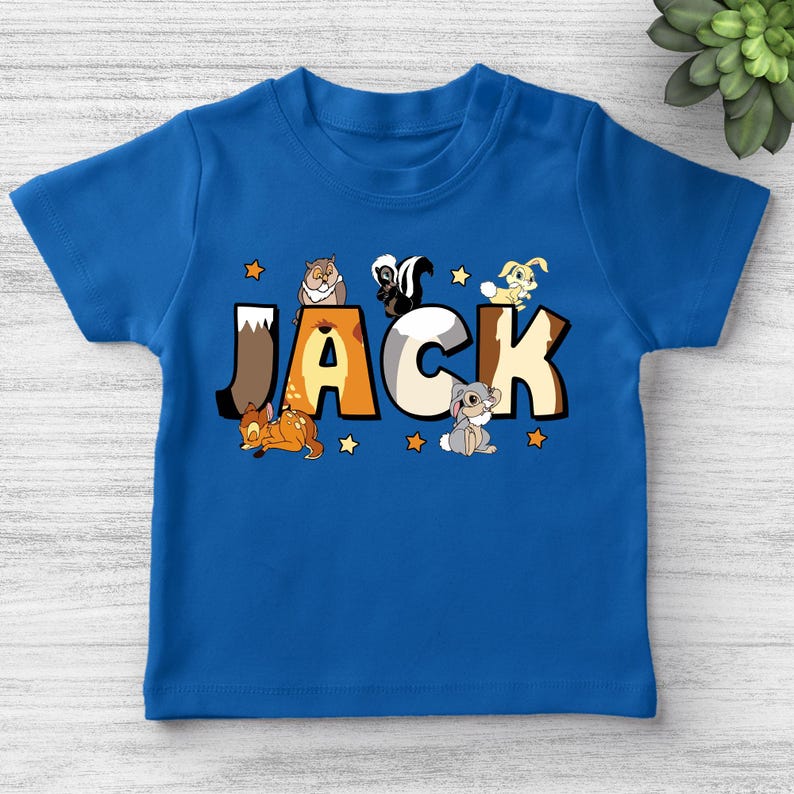 May include: A blue t-shirt with the name "JACK" printed across the front in large, colourful letters. The letters are decorated with cartoon animals, including a deer, skunk, rabbit, and hamster. Small yellow stars are scattered around the letters.