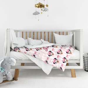 May include: A white crib with wooden slats containing a blanket featuring a repeating pattern of Minnie Mouse heads with pink bows and pink stripes. The blanket includes a personalized name in cursive script. A gray plush elephant sits nearby.
