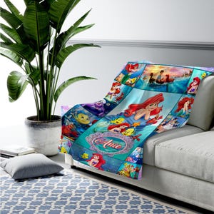 May include: A soft throw blanket featuring a collage of scenes and characters from The Little Mermaid, including Ariel and Flounder. The design includes a central decorative oval frame with the name Anie printed in a cursive red font.