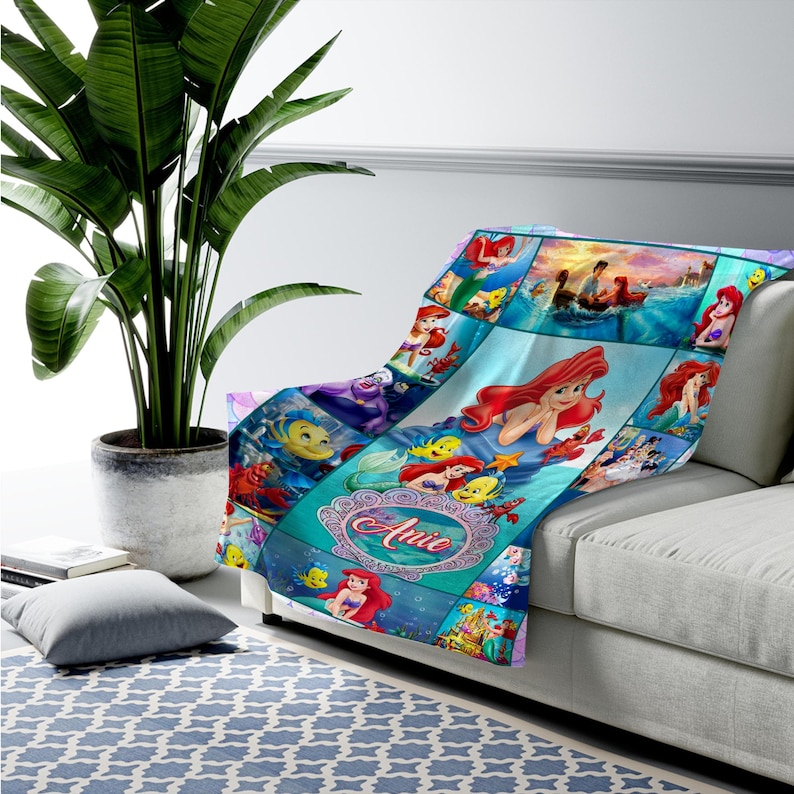 May include: A soft throw blanket featuring a collage of scenes and characters from The Little Mermaid, including Ariel and Flounder. The design includes a central decorative oval frame with the name Anie printed in a cursive red font.