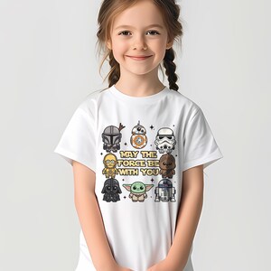 May include: White t-shirt featuring cartoon illustrations of Star Wars characters, including Darth Vader, a Stormtrooper, and Baby Yoda. The phrase "MAY THE FORCE BE WITH YOU" is printed below the characters. The shirt is worn by a young girl.