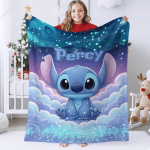 Puede incluir: Una manta rectangular de forro polar con un personaje alien&iacute;gena azul sentado entre nubes blancas sobre un cielo nocturno estrellado de color morado y azul. El nombre Percy est&aacute; impreso en letras azules grandes sobre el personaje.
