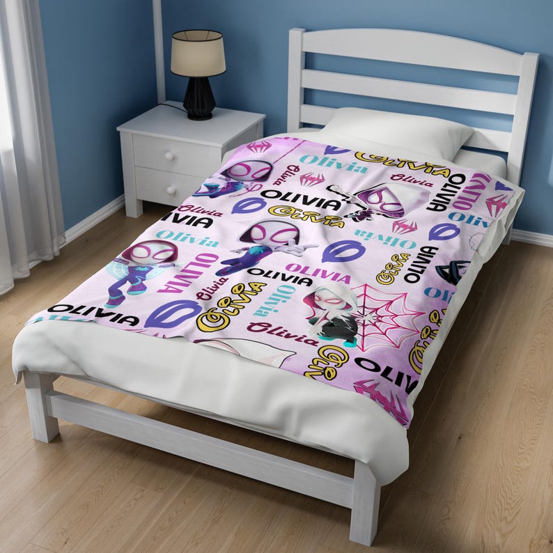 May include: A light pink blanket featuring a repeating pattern of cartoon characters in white, purple, and black superhero suits, red spider web graphics, and the name Olivia printed in various fonts, colors, and sizes across the fabric surface.
