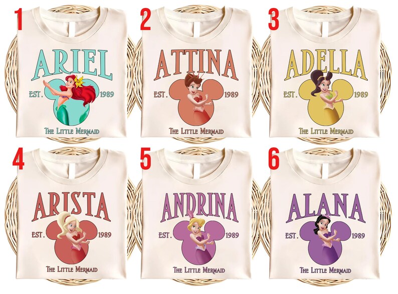 Puede incluir: Seis camisetas color crema con nombres e ilustraciones de personajes de "La Sirenita". Cada camiseta muestra un nombre, el a&ntilde;o 1989 y un dise&ntilde;o en forma de Mickey Mouse con una imagen de personaje.