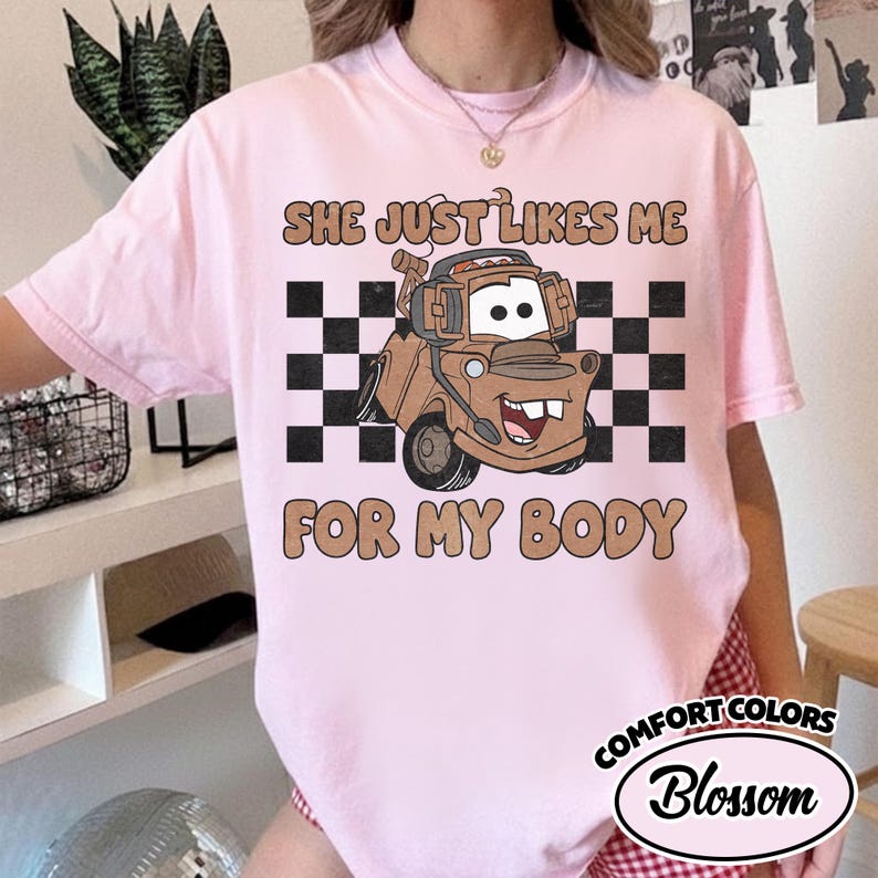 May include: A light pink t-shirt featuring a graphic of a brown tow truck character wearing headphones. The design includes a black and white checkered background and the text "SHE JUST LIKES ME FOR MY BODY" in a tan, rounded, bold font.