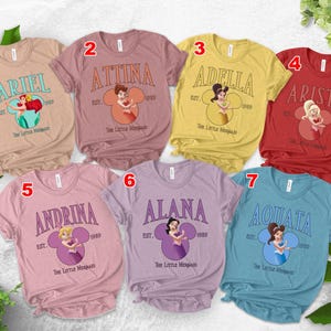 Puede incluir: Siete camisetas coloridas con personajes de La Sirenita de Disney. Cada camiseta muestra el nombre de un personaje en una fuente vintage, con una ilustraci&oacute;n del personaje a juego dentro de un dise&ntilde;o en forma de Mickey Mouse. Las camisetas son de varios colores.