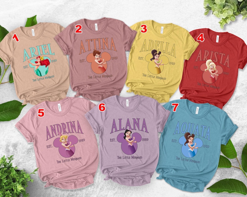 Puede incluir: Siete camisetas coloridas con personajes de La Sirenita de Disney. Cada camiseta muestra el nombre de un personaje en una fuente vintage, con una ilustraci&oacute;n del personaje a juego dentro de un dise&ntilde;o en forma de Mickey Mouse. Las camisetas son de varios colores.