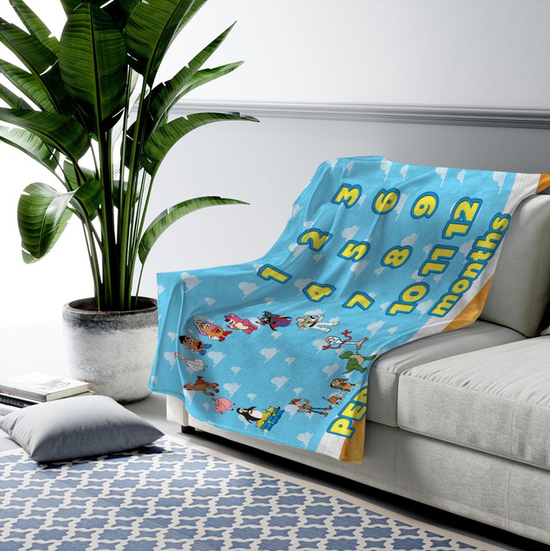May include: A light blue fleece blanket featuring a repeating white cloud pattern and various animated toy characters arranged in a circle. The blanket includes yellow numbers from 1 to 12 and the word months printed in a bold yellow font.