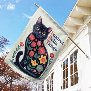 May include: A decorative outdoor flag featuring a black cat with floral patterns. The cat's body is adorned with red, yellow, and pink flowers. The flag also displays the text "Foxtrot Delta Tango" in red and white.