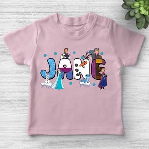 May include: A light pink t-shirt featuring the name "JANE" in colorful, cartoon-style letters. Each letter is decorated with characters from a popular animated movie, including a snowman, a princess, and other figures. Blue snowflake accents are scattered around the name.
