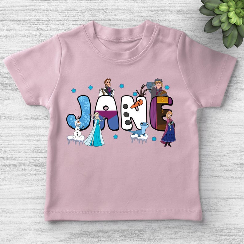 May include: A light pink t-shirt featuring the name "JANE" in colorful, cartoon-style letters. Each letter is decorated with characters from a popular animated movie, including a snowman, a princess, and other figures. Blue snowflake accents are scattered around the name.