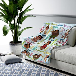 May include: A light blue throw blanket featuring a repeating pattern of Moana characters, including Moana, Maui, Pua the pig, and Heihei the rooster, interspersed with the text Moana in various colorful fonts and styles across the fabric surface.
