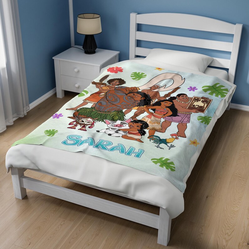 K&ouml;nnte beinhalten: Eine hellblaue Fleecedecke mit einem farbenfrohen Moana-Grafikdruck, der Charaktere wie Moana, Maui, Pua und Heihei zeigt, akzentuiert durch tropische Blumen und gr&uuml;ne Bl&auml;tter, mit dem Namen Sarah in gro&szlig;en blauen Buchstaben aufgedruckt.