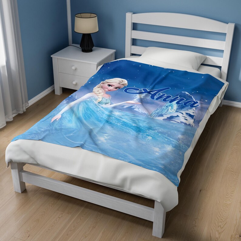 May include: A blue fleece blanket featuring a graphic print of a blonde character in a light blue dress, with the name Anna written in a dark blue script font. The blanket displays a snowy landscape with ice formations and a starry night sky.
