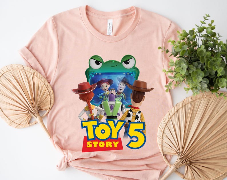 May include: A light pink short-sleeved t-shirt featuring a graphic print of characters from Toy Story, including Woody, Jessie, and Buzz Lightyear, positioned in front of a green frog-shaped frame with the text Toy Story 5 printed below.