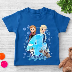 Pode incluir: Uma camiseta azul com um design gr&aacute;fico com o n&uacute;mero 4, duas personagens femininas animadas, um boneco de neve e uma rena. O nome "Sophia" est&aacute; escrito em it&aacute;lico abaixo do n&uacute;mero. Flocos de neve est&atilde;o espalhados ao redor do design.
