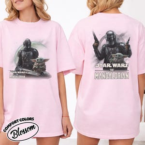 Puede incluir: Camiseta rosa claro con un dise&ntilde;o de Star Wars. La parte delantera muestra a The Mandalorian y Grogu con el texto "STAR WARS THE MANDALORIAN". La parte trasera muestra a The Mandalorian con armas, Grogu y el mismo texto.