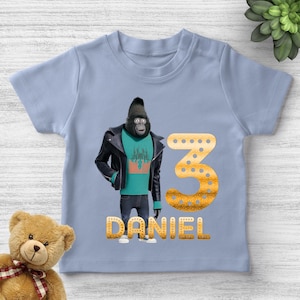 May include: Light blue toddler t-shirt featuring a cartoon gorilla character wearing a black jacket and jeans. The number 3 and the name "DANIEL" are printed in gold glitter font. A teddy bear is visible in the lower left corner.