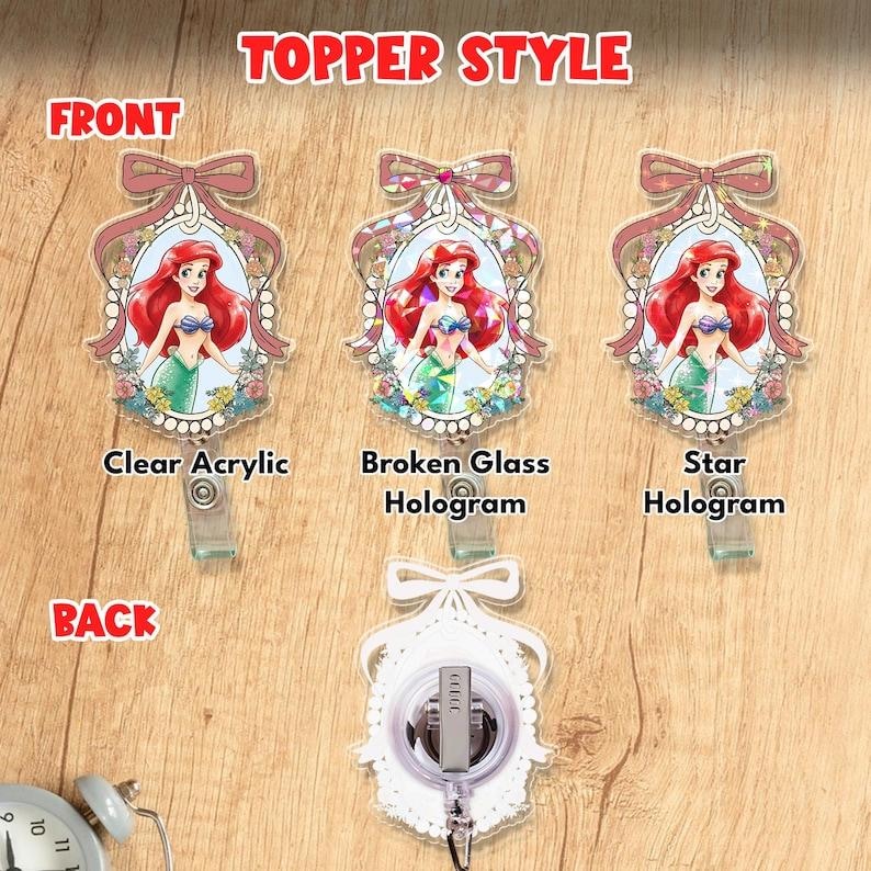 Puede incluir: Tres portatarjetas con Ariel la sirenita en un marco ovalado con lazo. Las etiquetas identifican los estilos como Clear Acrylic, Broken Glass Hologram y Star Hologram. Un cuarto muestra la parte trasera con clip. El texto dice TOPPER STYLE.