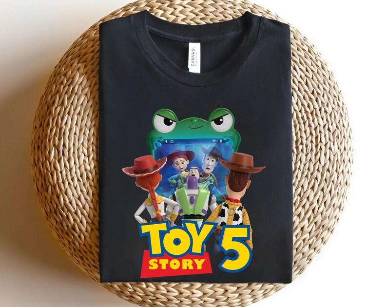 May include: A black short-sleeved t-shirt featuring a graphic print of characters from Toy Story looking at a screen framed by a green frog. Below the characters, the text reads Toy Story 5 in large yellow and red letters. The shirt has a crew neckline.