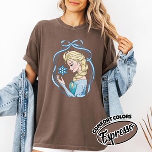 May include: A brown t-shirt featuring a cartoon illustration of a blonde woman with a braid, holding a snowflake. The illustration is framed by a blue ribbon design. The text "COMFORT COLORS Espresso" is printed on the bottom right.