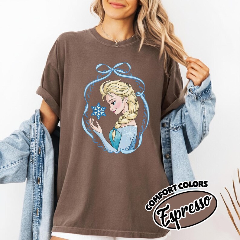 May include: A brown t-shirt featuring a cartoon illustration of a blonde woman with a braid, holding a snowflake. The illustration is framed by a blue ribbon design. The text "COMFORT COLORS Espresso" is printed on the bottom right.