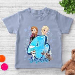 Pode incluir: Uma camiseta azul clara para crian&ccedil;as com um design do Frozen. O design inclui as personagens Anna e Elsa, o n&uacute;mero 4 e o nome "Sophia" em it&aacute;lico. Tamb&eacute;m est&atilde;o presentes flocos de neve e outros personagens do filme.