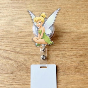 May include: A badge reel featuring a cartoon image of Tinkerbell in a seated pose, wearing a green dress and with translucent wings. The reel has a clear plastic strap and a white rectangular card holder.