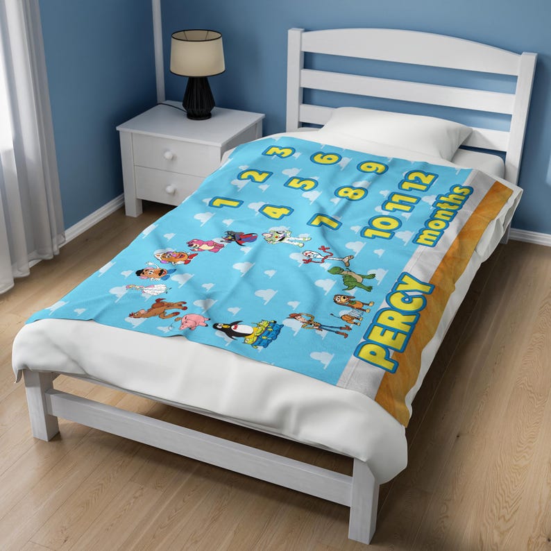 May include: A light blue fleece blanket featuring a repeating white cloud pattern, a yellow numerical sequence from 1 to 12, the word months, and various cartoon character illustrations. The name PERCY is printed in large yellow letters on the side.