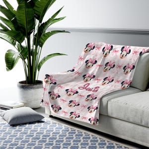 May include: A soft throw blanket featuring a repeating pattern of Minnie Mouse faces with pink bows on a light pink background. The name Julia is printed in a cursive script font in the center of the blanket, which is draped over a sofa.