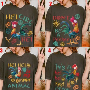 Op de afbeelding: Vier donkergrijze t-shirts met hanenafbeeldingen en tekst. Het eerste shirt zegt "HEI GIRL HEI". De tweede zegt "DON'T BE A Peeker". De derde zegt "HEI HEI IS MY SPIRIT ANIMAL". De vierde zegt "It's a hei hei Kind Of day".