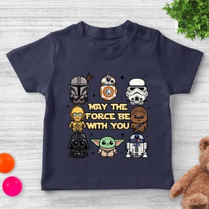 May include: Navy blue toddler t-shirt featuring cartoon illustrations of Star Wars characters, including The Mandalorian, BB-8, and Darth Vader. The text "MAY THE FORCE BE WITH YOU" is printed in the center of the shirt.