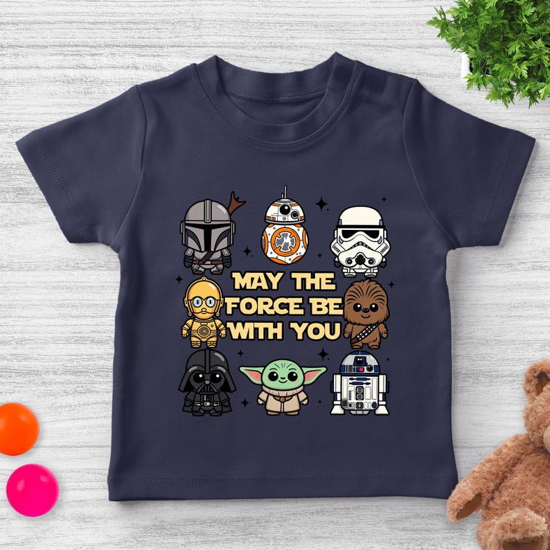 May include: Navy blue toddler t-shirt featuring cartoon illustrations of Star Wars characters, including The Mandalorian, BB-8, and Darth Vader. The text "MAY THE FORCE BE WITH YOU" is printed in the center of the shirt.