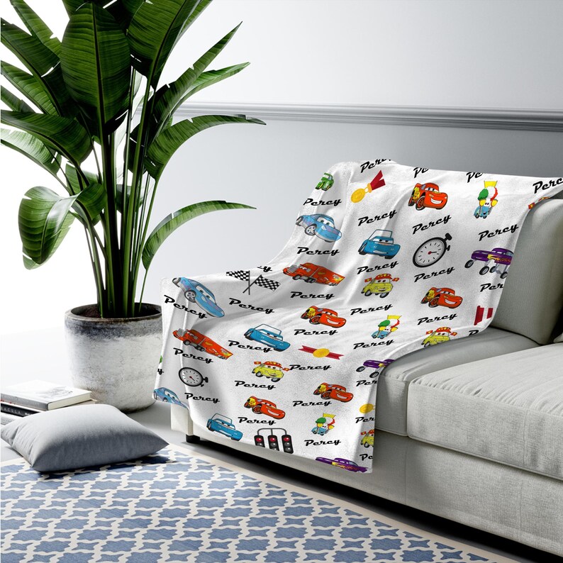 May include: A white throw blanket featuring a repeating pattern of cartoon race cars, checkered flags, stoplights, stopwatches, and gold medals, interspersed with the name Percy written in a black cursive script across the fabric surface.