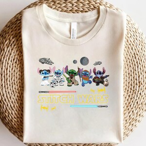 May include: Cream-colored t-shirt featuring a graphic of several cartoon characters in various costumes, with the text "Stitch Wars" in yellow and the image of two lightsabers. The shirt is made of a soft material.