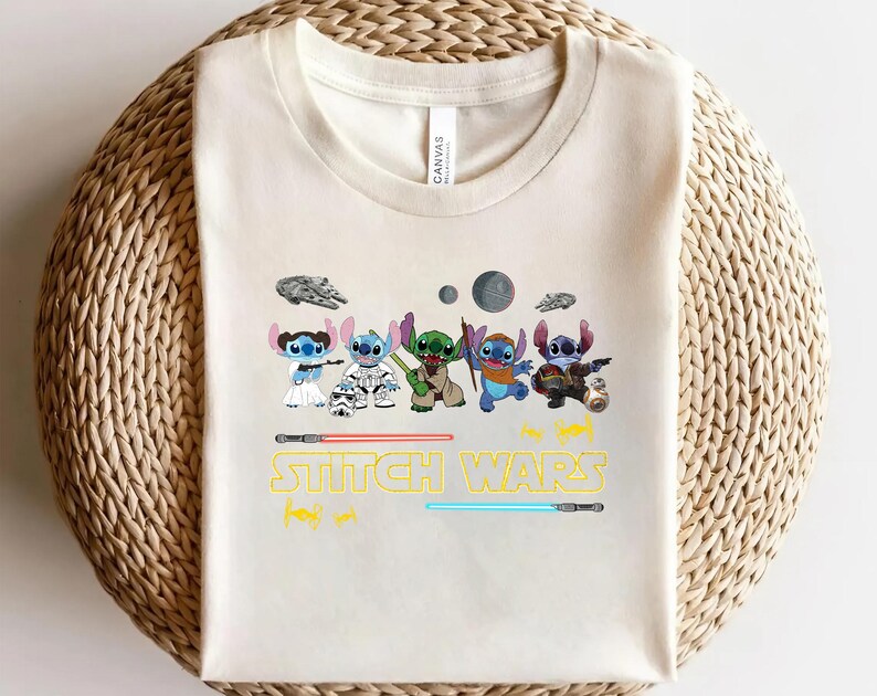 May include: Cream-colored t-shirt featuring a graphic of several cartoon characters in various costumes, with the text "Stitch Wars" in yellow and the image of two lightsabers. The shirt is made of a soft material.