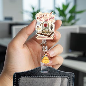 May include: A clear badge reel featuring a Beauty and the Beast theme. The design includes a rose in a glass dome, the text "BELLE'S BOOK CLUB", and an image of Belle in a yellow dress. The reel also has the text "TALE AS OLD AS TIME".