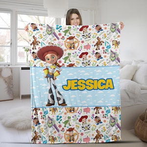 May include: A white fleece blanket featuring a repeating pattern of Toy Story characters including Jessie, Woody, Buzz Lightyear, Rex, and Mr. Potato Head, with a central light blue horizontal band displaying the name JESSICA in yellow text.